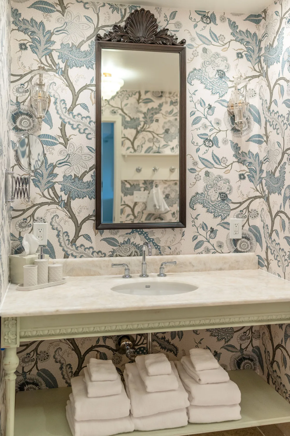 A calming and sophisticated bathroom scene featuring an ornate mirror, decorative wallpaper, a modern sink and faucet, and towels neatly stacked. The elegant setting promotes relaxation and rejuvenation, with thoughtful details that evoke a sense of personal indulgence.