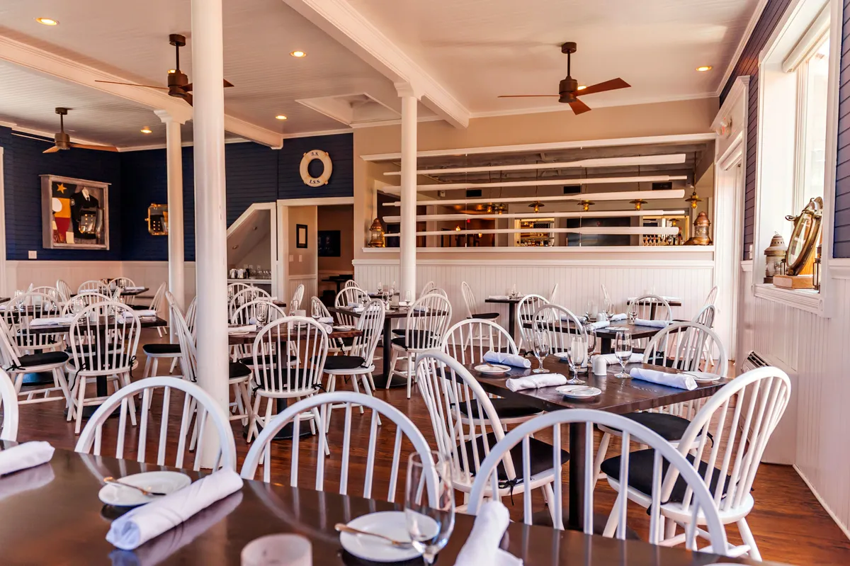 A stylish, casual dining area with white wooden chairs and dark tables set against decorative wall art, with a bar area visible in the background. The scene suggests a lively yet comfortable space for dining, socializing, and gathering with friends or family.