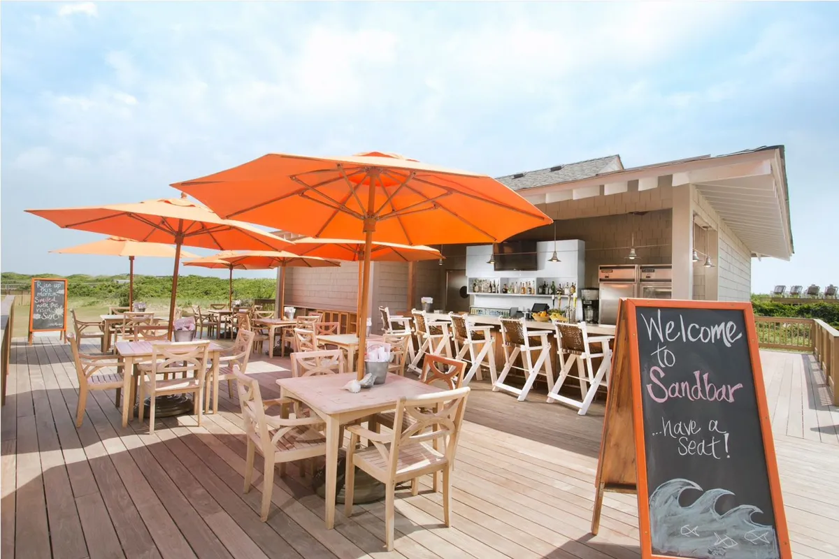 An inviting outdoor dining area with orange umbrellas, wooden tables, and a welcoming chalkboard sign. The space is designed for casual dining and socializing, with a relaxed, cheerful mood. The dining setup suggests convenience and kid-friendly options for families after a day in the sun.