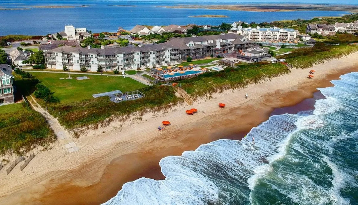 This image captures a sweeping view of the property’s beachfront setting, showcasing the inviting swimming pool, the hotel complex itself, and rows of umbrellas lining the sandy shore. The scene conveys opportunities for swimming, sunbathing, and relaxing moments by the water, with the dunes in the background hinting at the scenic vistas couples can enjoy together.