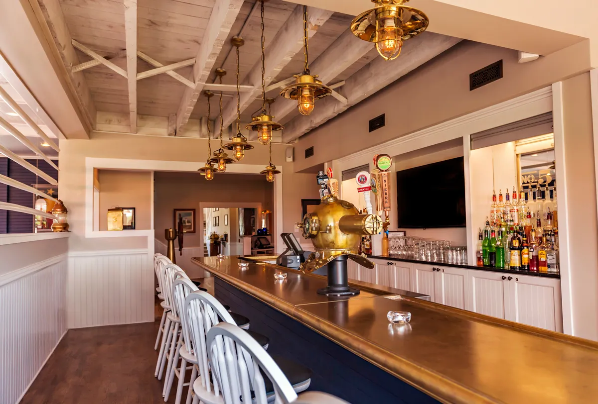 A lively bar area with a polished counter, stylish hanging lights, and shelves lined with bottles. Nautical décor and an inviting vibe make this space perfect for cocktails or socializing after a day of wellness and activity.