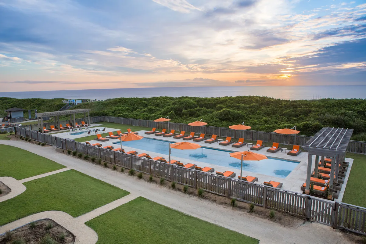 This image captures a sparkling outdoor swimming pool surrounded by lounge chairs and vibrant orange umbrellas, all set against a backdrop of the ocean at sunset. The tranquil atmosphere is enhanced by subtle hints of social gatherings and relaxation, with the warm evening light inviting guests to unwind.
