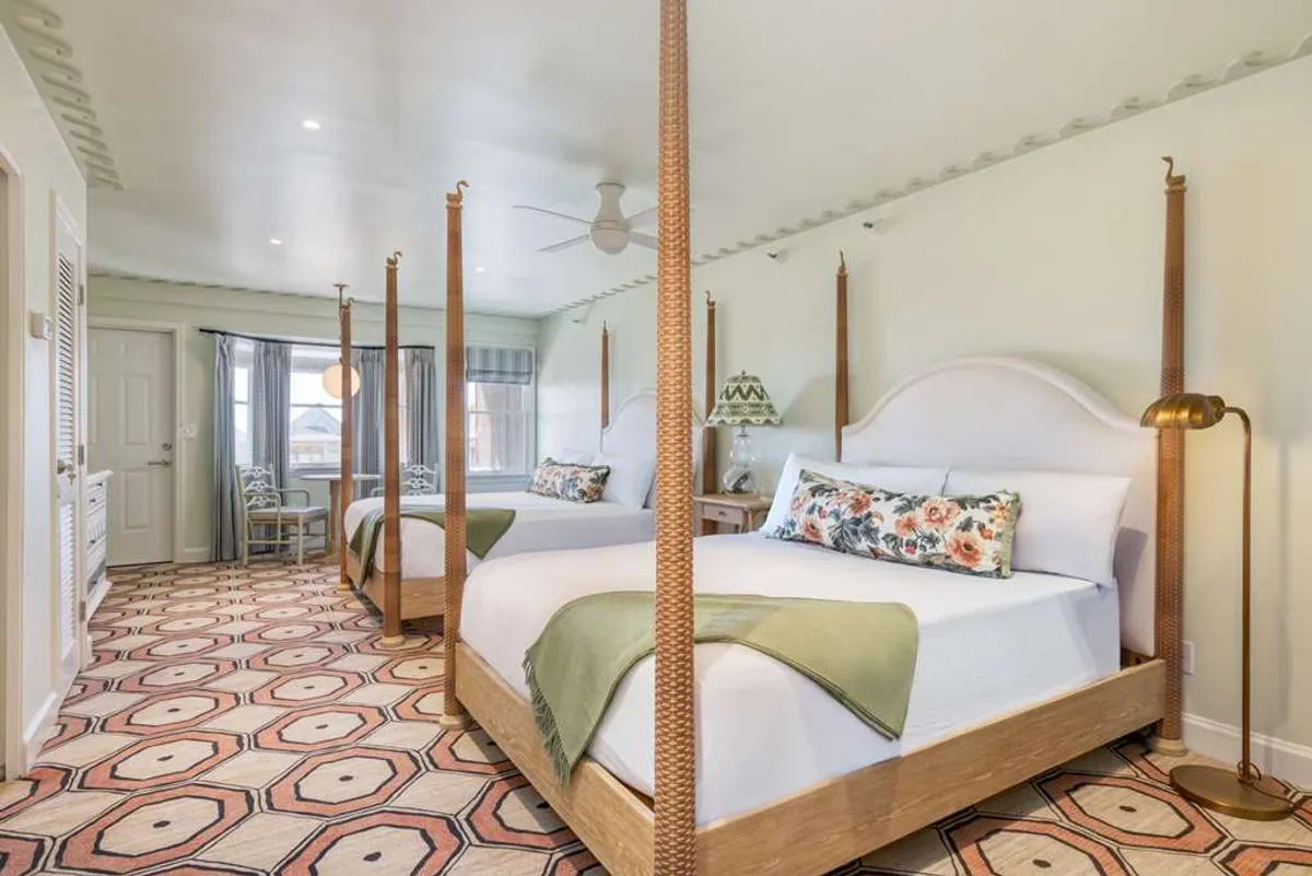 A beautifully decorated guestroom featuring two four-poster queen beds with colorful floral pillows, a geometric patterned rug, and a decorative lamp. Sunlight filters into the serene space, evoking comfort and understated elegance. The room's spaciousness invites relaxation and socializing, ideal for families or couples.