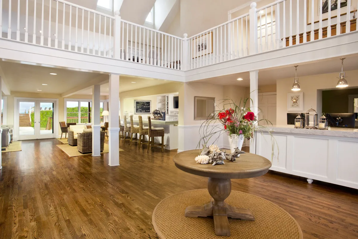A bright, spacious interior features a central round table adorned with a floral arrangement, large windows letting in natural light, and a modern open kitchen in the background. The open-plan layout signals a welcoming space ideal for gathering, relaxing, and enjoying flexible resort living.