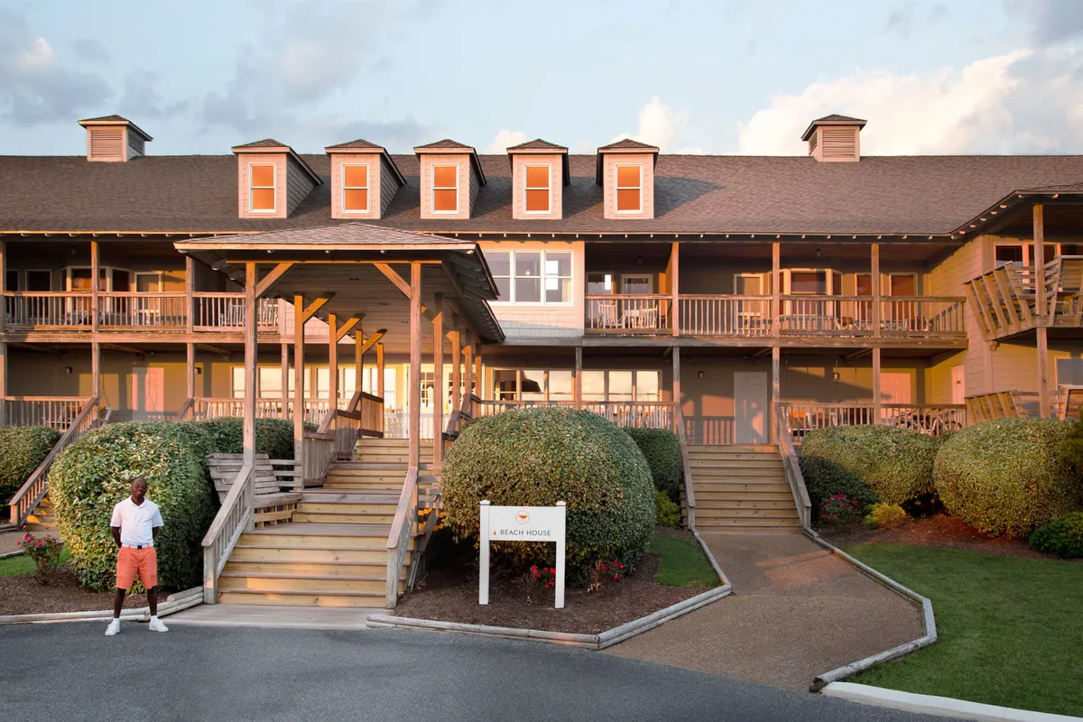 A charming exterior shot of the beach house, featuring stairs leading to a welcoming porch surrounded by landscaped bushes. The setting sun hints at the potential for cozy evenings spent together on the porch, as well as easy access to nearby beach trails.