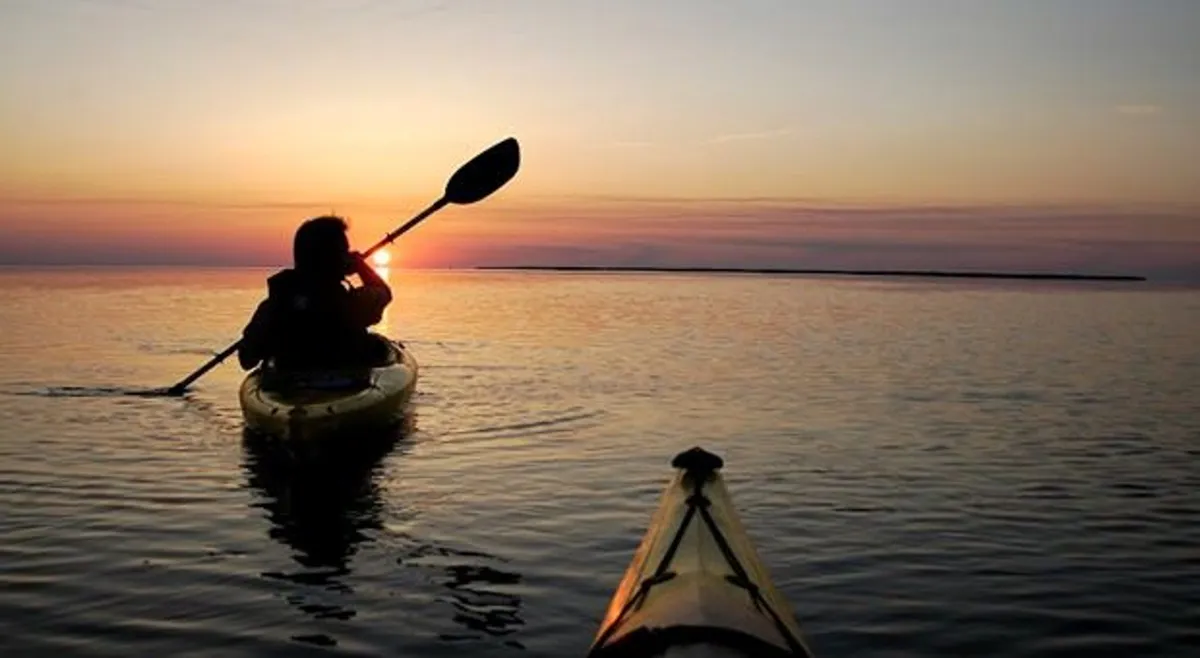 A tranquil sunset scene over calm water, with a kayaker paddling quietly in the foreground. The warm glow of the setting sun casts a romantic light across the water, evoking moments of reflection, connection, and the spirit of gentle adventure that couples can share.