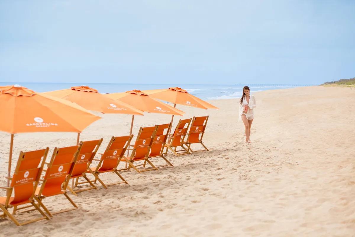 A tranquil beach scene featuring rows of beach chairs and vibrant orange umbrellas facing the ocean. A woman is seen walking towards the water, evoking a sense of freedom and relaxation. The overall ambiance is peaceful, with gentle waves and a soft sunset glow in the sky.