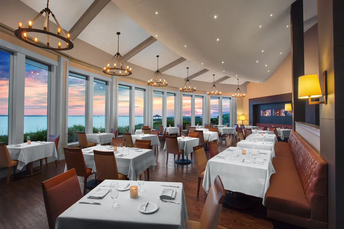 An elegant dining room featuring tables set with crisp white linens, comfortable leather seating, and warm chandelier lighting. Large windows showcase a sunset view, creating an inviting space for refined dining and special occasions.