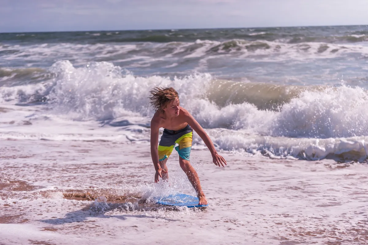 A joyful action shot of a young boy riding a boogie board on the waves, surrounded by soft sand and rolling surf. The lively mood reflects playful beach activities and family-friendly fun, with energy and movement at the heart of the image.