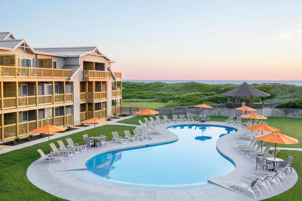 The photo features a vibrant outdoor swimming pool surrounded by orange umbrellas, sun loungers, and lush landscaped gardens. There are families and groups enjoying the cheerful atmosphere, swimming, sunbathing, or relaxing by the pool. The scenic balcony views and inviting pool area highlight the resort’s lively and communal spirit.
