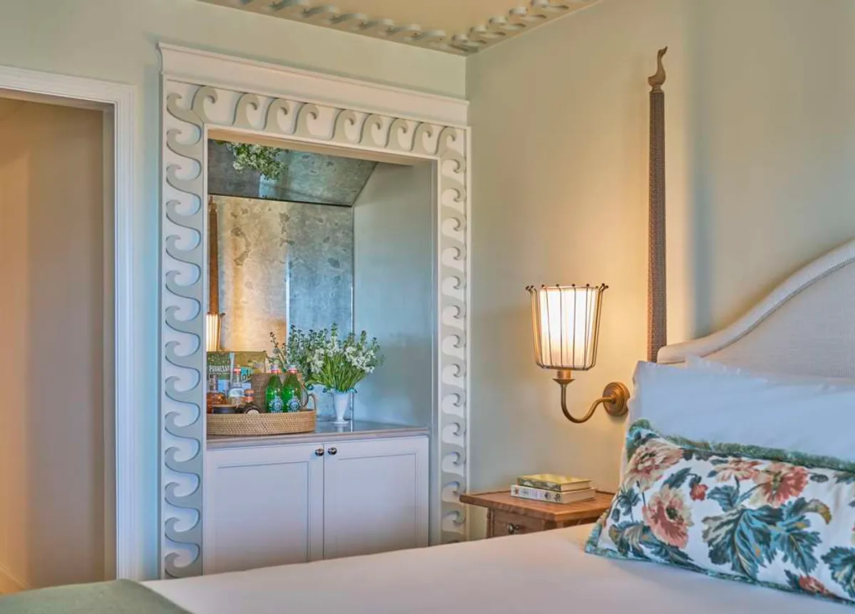 The image captures a serene corner of the studio guestroom at Sanderling Resort, featuring a decorative mirror, a cozy floral pillow, a vase of fresh flowers, and a warm wall sconce. Soft natural light fills the space, creating an inviting nook ideal for unwinding with a book or enjoying quiet moments. The decor balances understated elegance with comfort, highlighting the room’s peaceful atmosphere.
