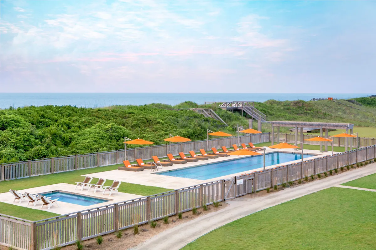 This image features an inviting outdoor swimming pool at Sanderling Resort, surrounded by comfortable lounge chairs and beach umbrellas. Lush greenery frames the pool area, creating a sense of tranquility and privacy. The scene is lively yet serene, with guests enjoying sunbathing, swimming, and socializing, all set against a backdrop of natural beauty.