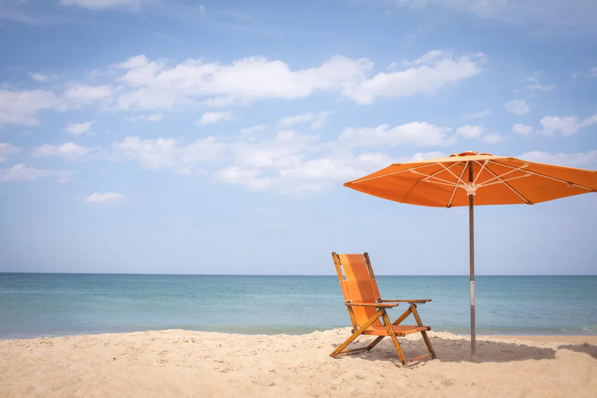 This image portrays a serene stretch of sandy beach lined with an orange beach chair and matching umbrella set against a calm ocean backdrop. The atmosphere is peaceful, inviting relaxation, sunbathing, or family activities like building sandcastles. The beach setting is open and family-friendly, ideal for children to play and enjoy the gentle surf.